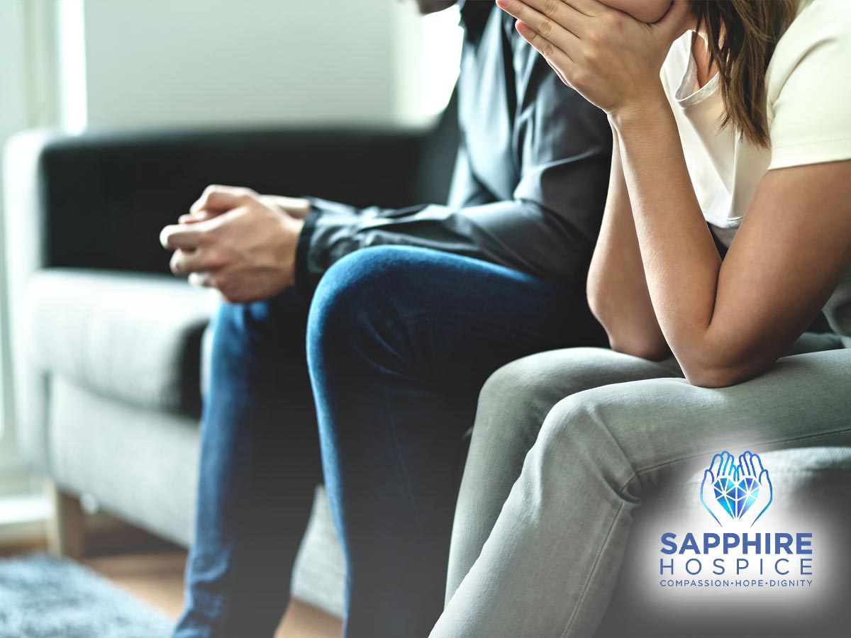 A couple in deep thought and grief sits on a couch, hands folded and heads bowed, symbolizing the process of bereavement